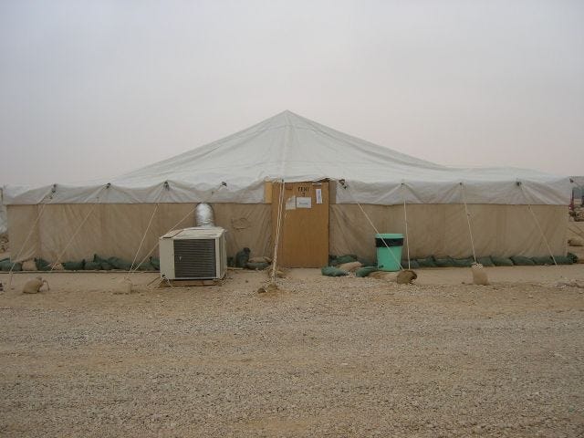 A photo of a large beige tent in a desert with a plywood door and a large air conditioning unit hooked up to the side.