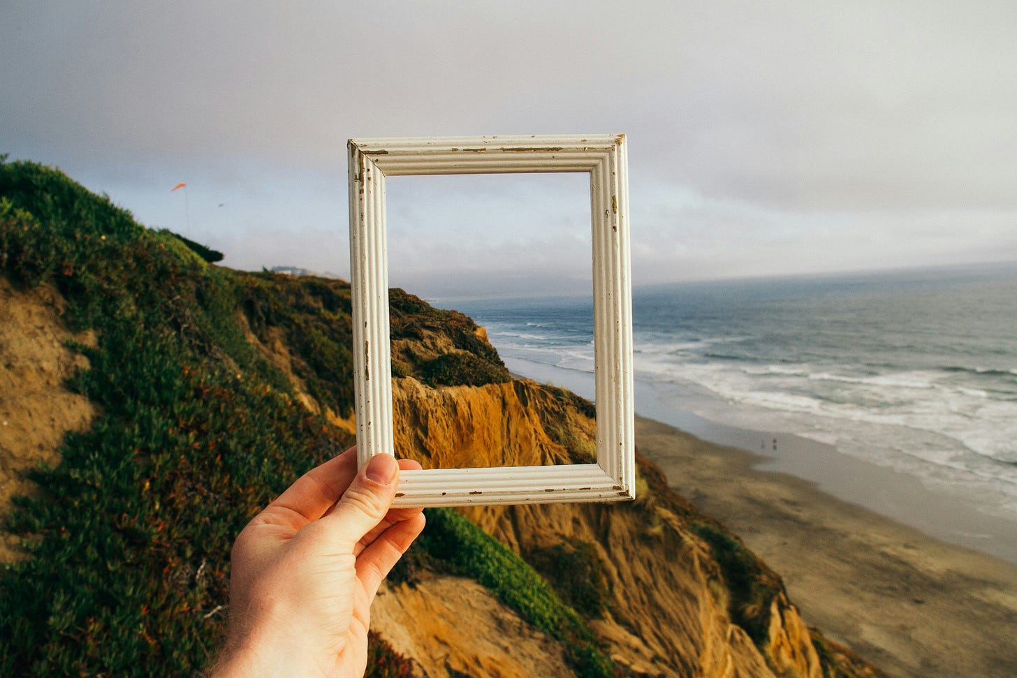 A left white hand holds a picture frame in front of a beautiful vista point of a beach.
