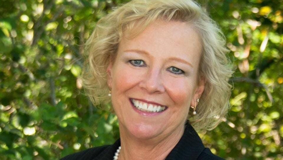 A woman with short blond hair, wearing a black blazer with an American flag lapel pin, poses for a portrait in front of a leafy tree. A woman with short blond hair, wearing a black blazer with an American flag lapel pin, poses for a portrait in front of a leafy tree.
