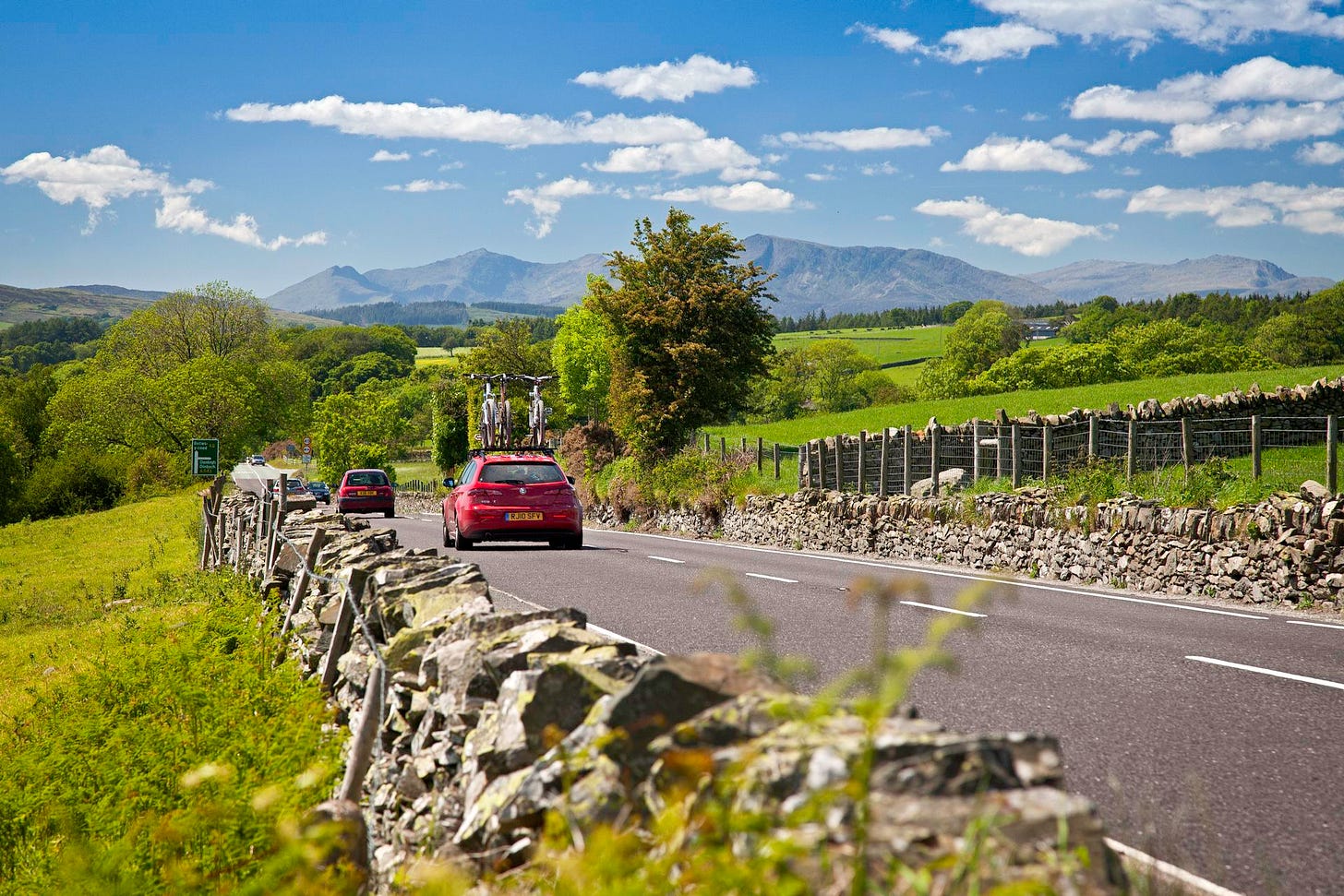 A bright red car carrying two bicycles on a roof rack drives along a paved country road bordered by rustic dry stone walls and lush green fields. In the background, a large, rugged mountain range stretches across the horizon under a sunny blue sky with scattered white clouds.