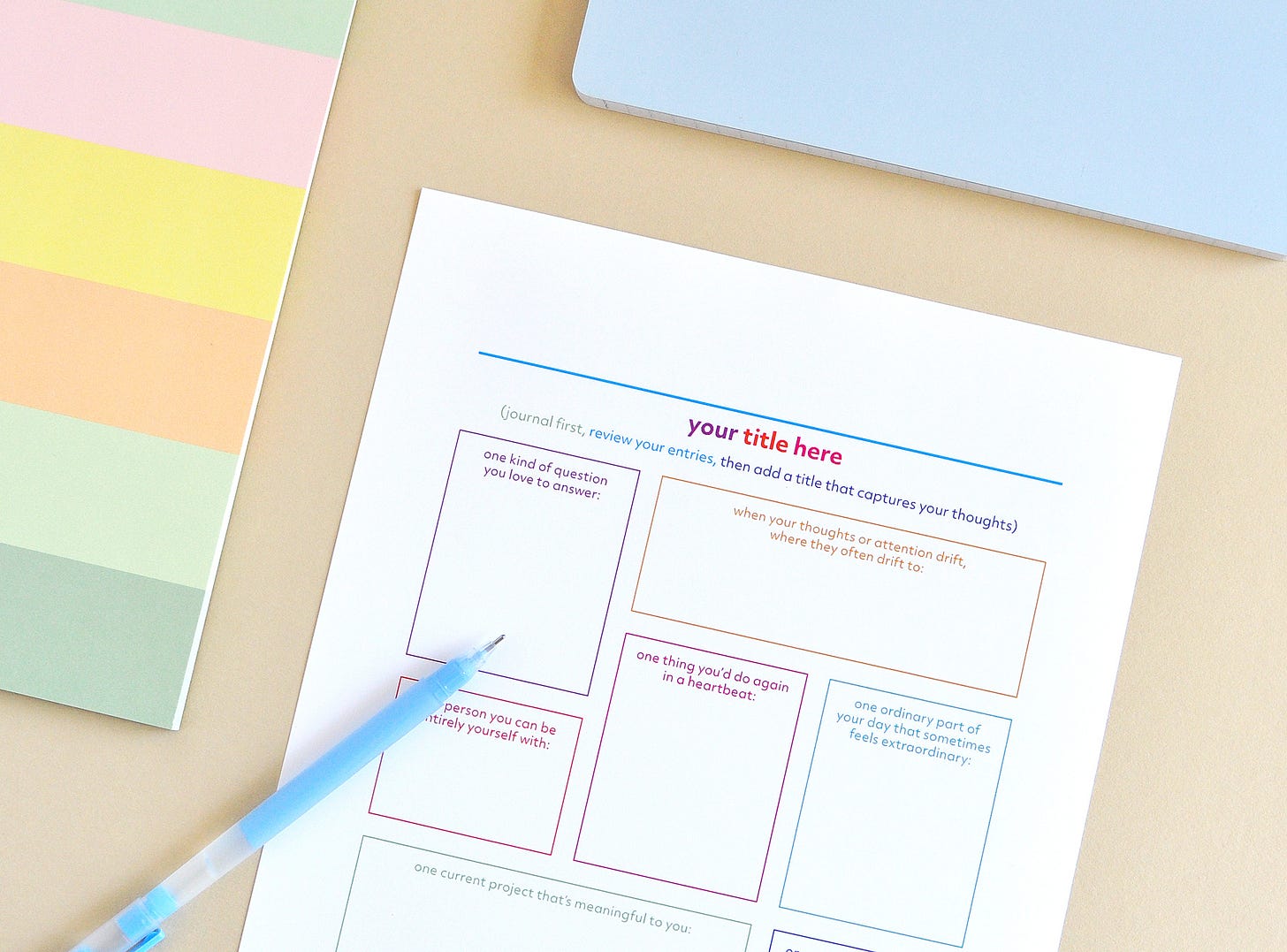 A colourful printable journal page filled with writing prompts and a space to write your own title after journaling. Shown with colourful notebooks and a blue pen on a warm beige background.