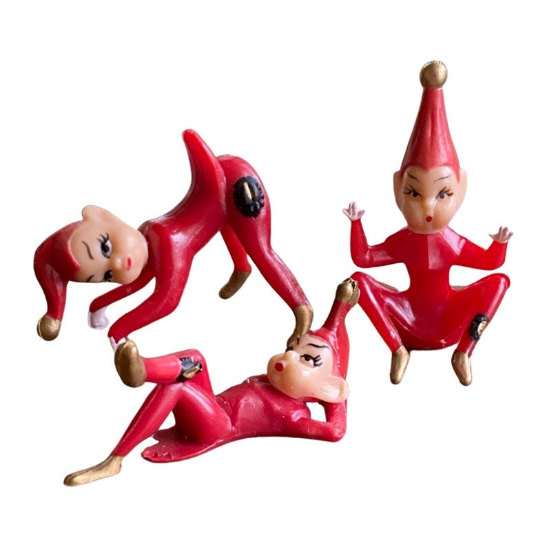 May include: Three red plastic Christmas elf figurines. The elves are wearing red hats and have painted faces. One elf is lying on its back, another is standing on its hands, and the third is sitting with its legs crossed. May include: Three red plastic Christmas elf figurines. The elves are wearing red hats and have painted faces. One elf is lying on its back, another is standing on its hands, and the third is sitting with its legs crossed.