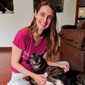 A imagem mostra uma mulher sentada no chão de uma sala, em um ambiente interno e bem iluminado. Ela sorri diretamente para a câmera, transmitindo uma expressão de alegria e tranquilidade. A mulher tem pele clara, cabelos longos e castanhos, soltos, e veste uma camiseta rosa e calça clara. Sobre suas pernas está um cachorro de porte médio, com pelagem curta nas cores preta e marrom. O cachorro também parece relaxado, com a boca levemente aberta, como se estivesse sorrindo, enquanto recebe carinho da mulher. Ao fundo, é possível ver um sofá marrom e parte da decoração da casa, criando uma sensação de ambiente acolhedor e familiar. A imagem mostra uma mulher sentada no chão de uma sala, em um ambiente interno e bem iluminado. Ela sorri diretamente para a câmera, transmitindo uma expressão de alegria e tranquilidade. A mulher tem pele clara, cabelos longos e castanhos, soltos, e veste uma camiseta rosa e calça clara. Sobre suas pernas está um cachorro de porte médio, com pelagem curta nas cores preta e marrom. O cachorro também parece relaxado, com a boca levemente aberta, como se estivesse sorrindo, enquanto recebe carinho da mulher. Ao fundo, é possível ver um sofá marrom e parte da decoração da casa, criando uma sensação de ambiente acolhedor e familiar.