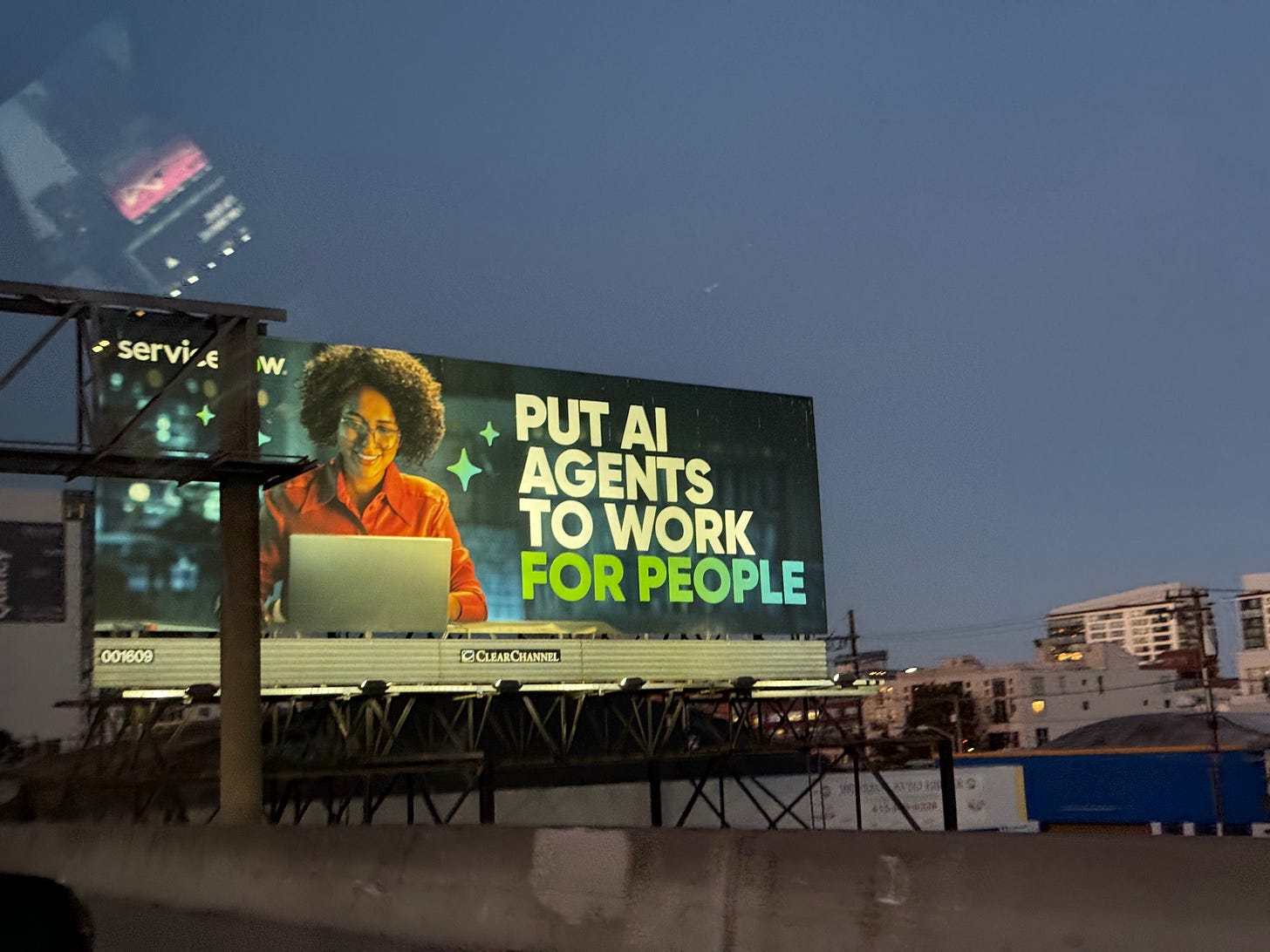 A roadside billboard at dusk shows a smiling person with curly hair wearing glasses and a red shirt, working on a laptop. The text on the billboard reads, “PUT AI AGENTS TO WORK FOR PEOPLE.”