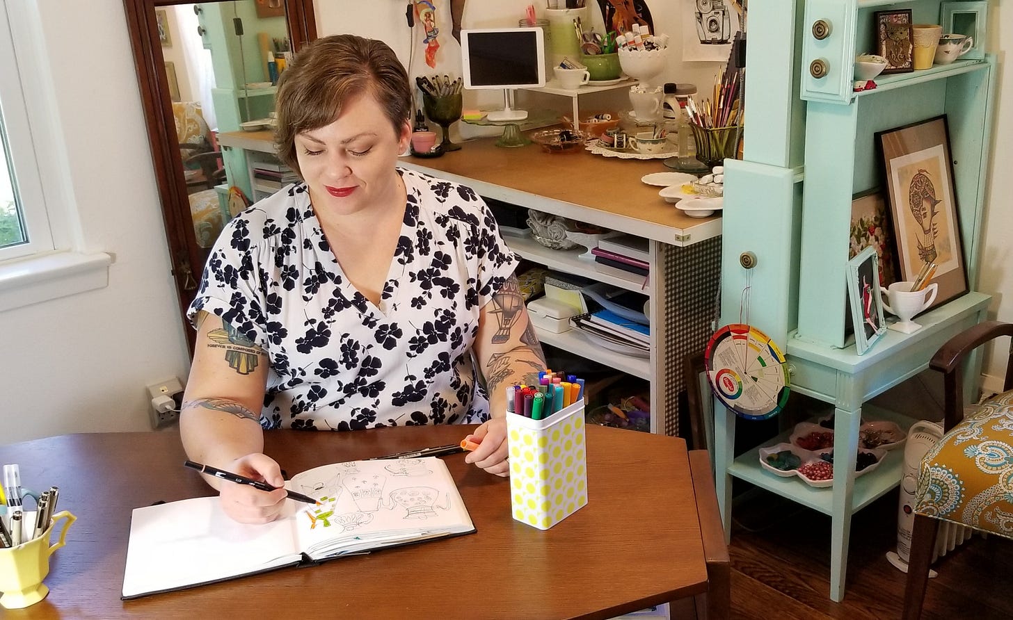 Artist Beth Hempton sitting at a desk in an art studio, coloring in a sketchbook with a marker. Artist Beth Hempton sitting at a desk in an art studio, coloring in a sketchbook with a marker.