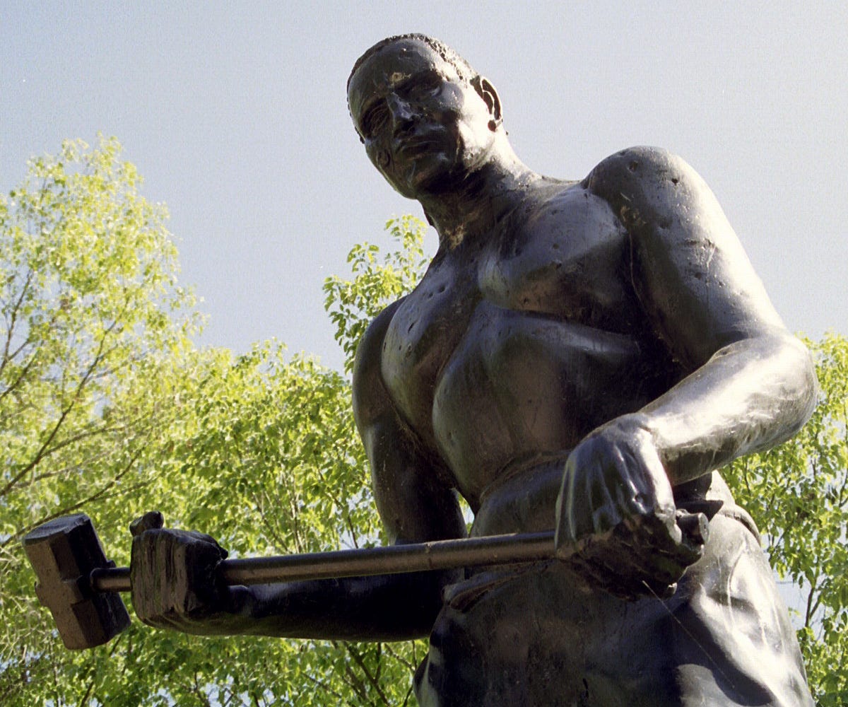 A bronze statue of John Henry photographed from below against sky and trees. He is shirtless and muscular, gripping a sledgehammer across his body with both hands. A bronze statue of John Henry photographed from below against sky and trees. He is shirtless and muscular, gripping a sledgehammer across his body with both hands.