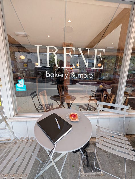 A table outside a bakery called Irene, with a black journal, pen, coffee in a glass cup and a postcard with pop art design.