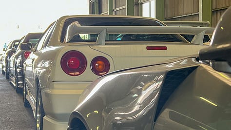 A collection of photos showing dozens of white, blue and green Nissan R34 Skyline GT-R sports cars crammed into a dimly-lit warehouse with gray walls and green support beams with a gravel floor.
