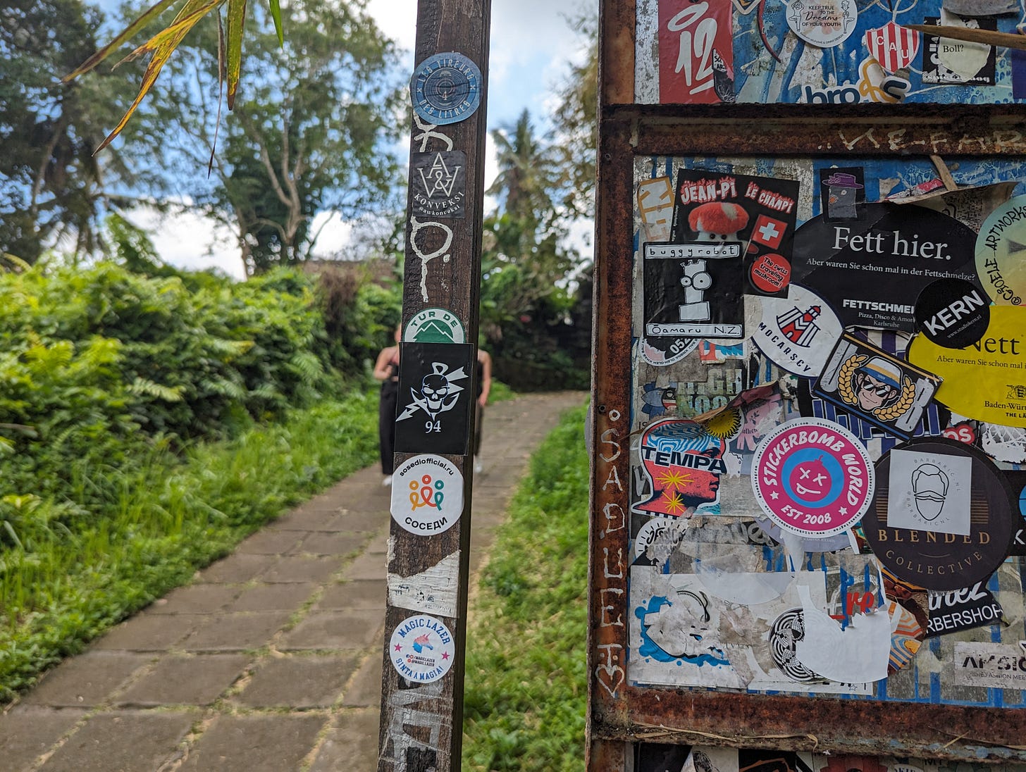 A weathered post and gate covered in various stickers, including a prominent 'Stickerbomb World' sticker, set against a lush green outdoor background with a stone pathway leading into the distance.