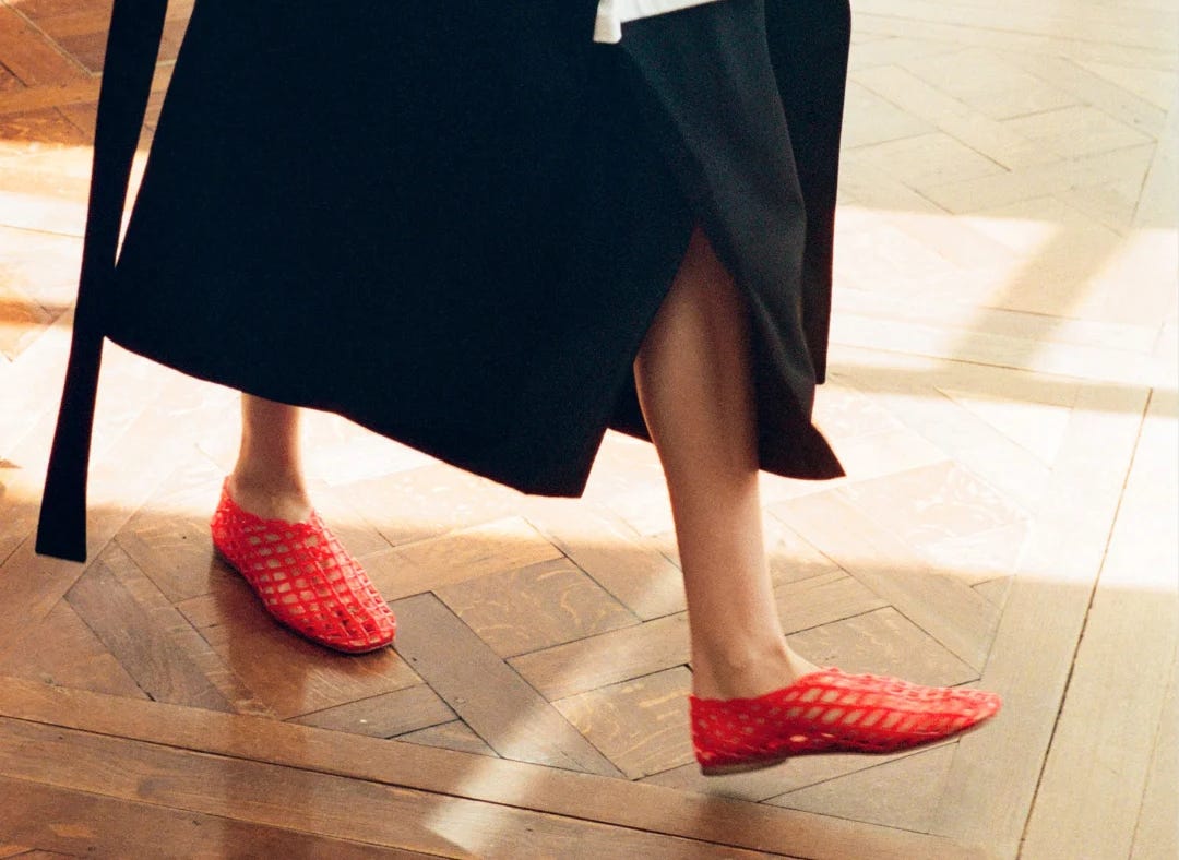 A closeup image detailing a pair of laticed jelly flats worn by a woman as she walks the runway, a trace of her black coat visible toward the top of the image.