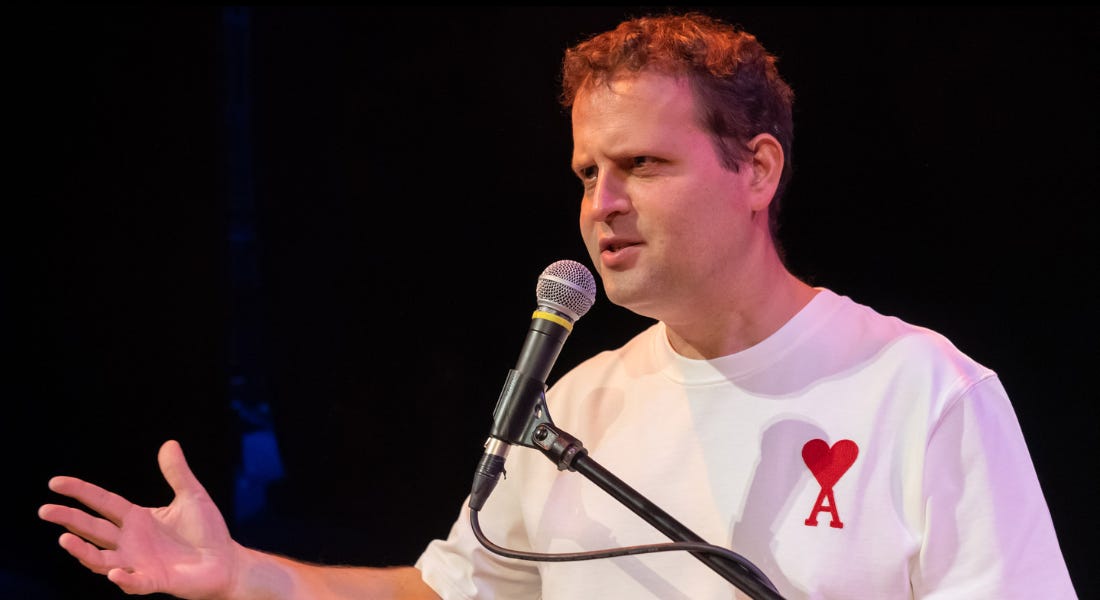 Adam Kay wearing a white t-shirt with a red 'A' on it standing in front of a black microphone with a black backdrop behind him
