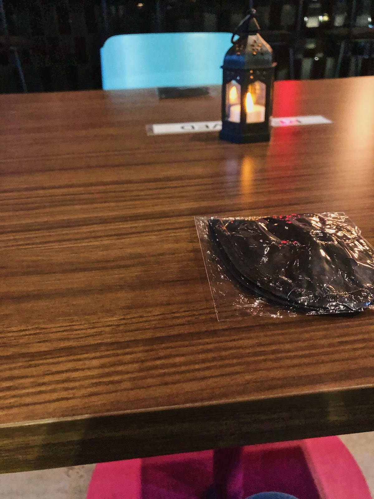 Picture of a candlelit table with a plastic wrapped new black blindfold on top of the table and a blue chair on the opposite end of the table.