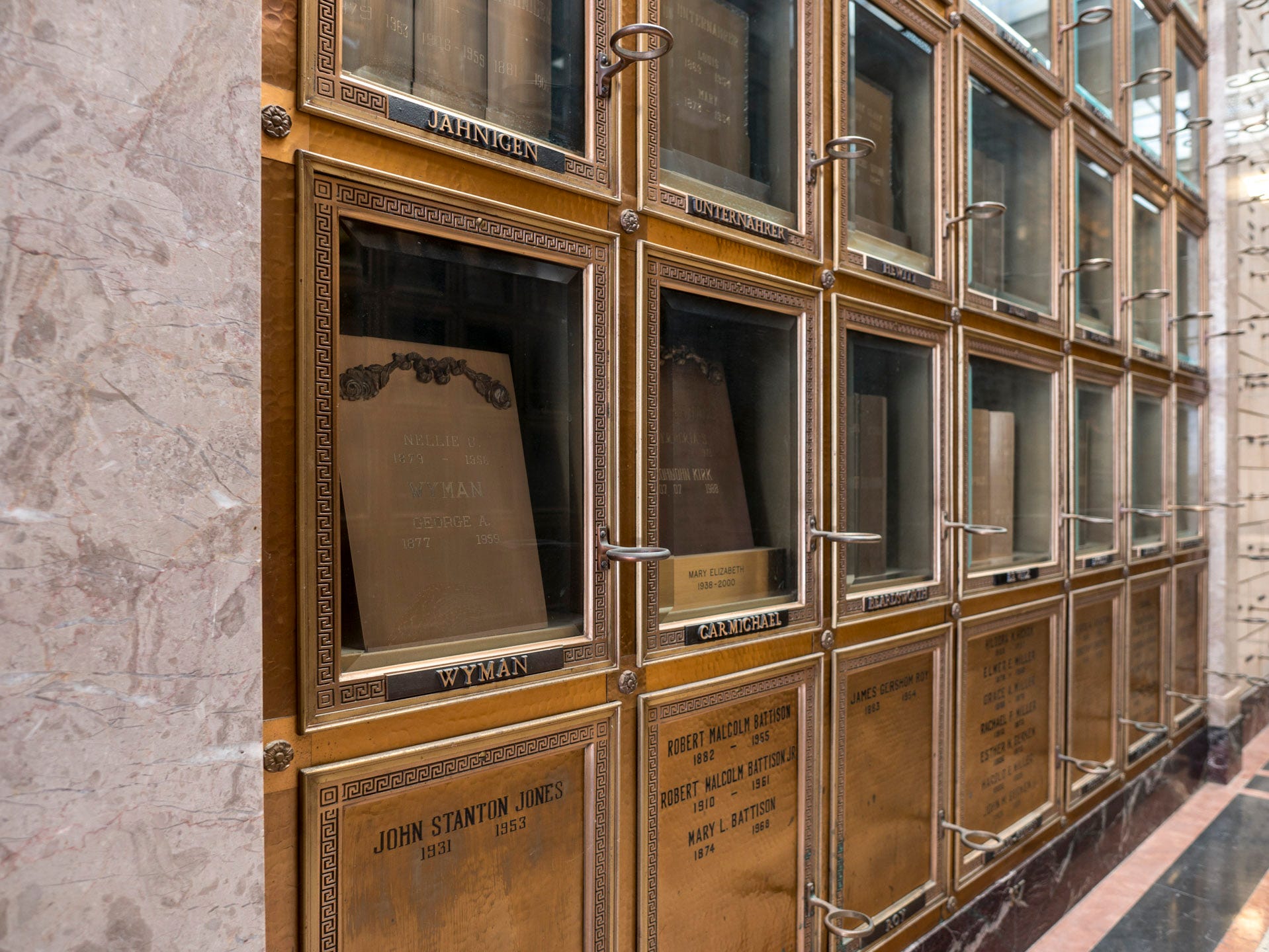 The remains of George A Wyman and his wife Nellie interred inside a mausoleum. The remains reside behind a glass door, part of a wall of similar interments. The doors and trim are finished in bronze. Small votive holders are attached to each grave.