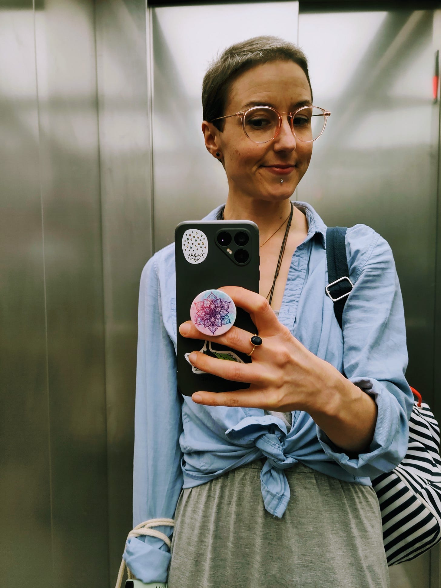 The photo shows a selfie of the author, a white woman with short brown hair, in the elevator. She is wearing a long grey dress with a blue shirt, glasses, and carries a backpack.