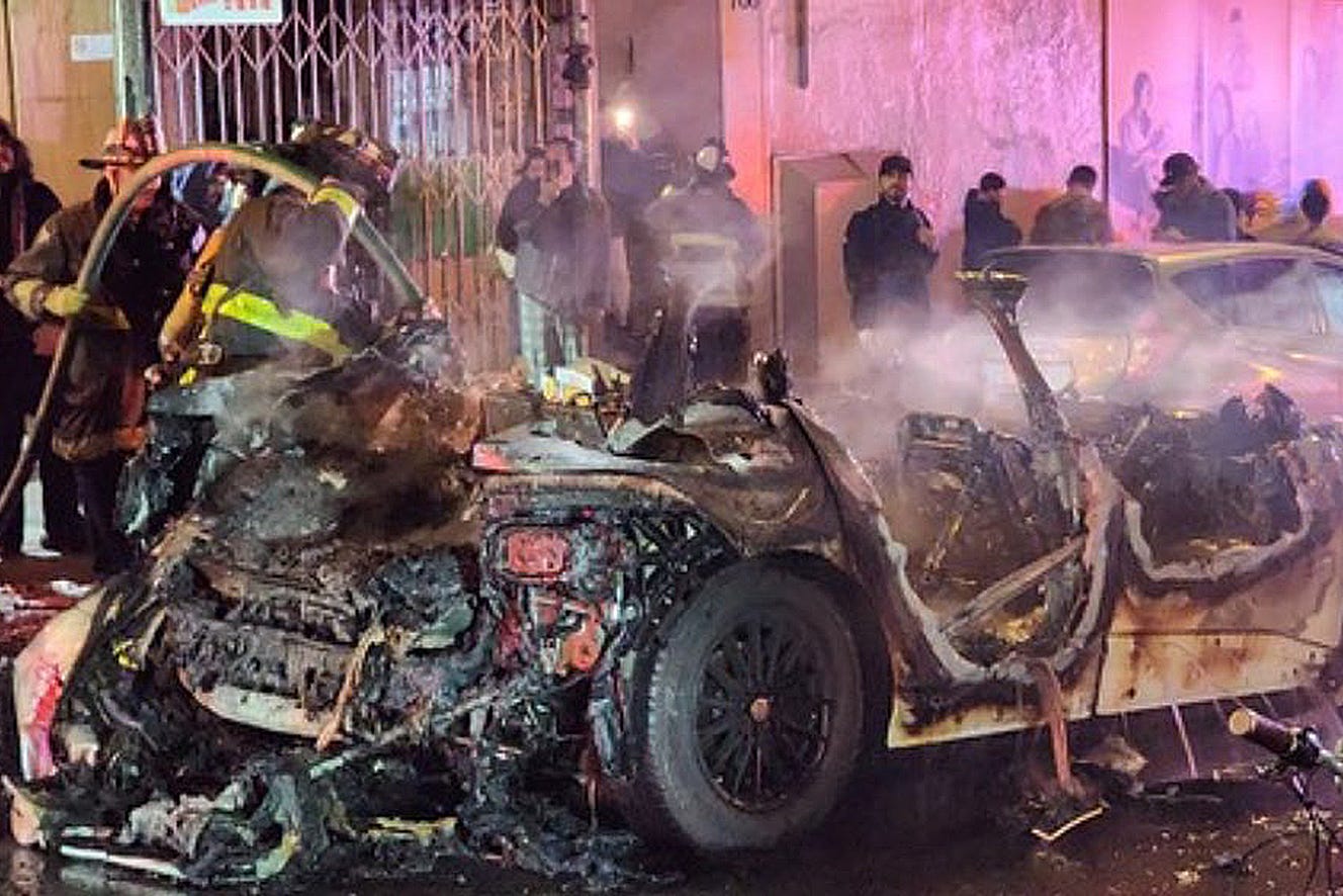 A firefighter hoses down a charred car wreck at night, with onlookers behind barriers. A firefighter hoses down a charred car wreck at night, with onlookers behind barriers.