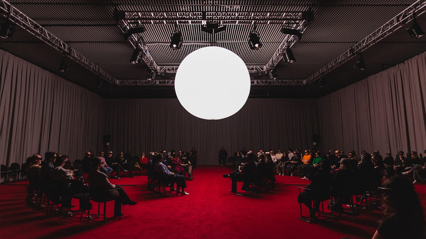 A large curtained performance space with a bright red carpet. Audience sit in concentric circles wearing mixed reality headsets. At the center of the room hangs a glowing white orb-like light fixture. A large curtained performance space with a bright red carpet. Audience sit in concentric circles wearing mixed reality headsets. At the center of the room hangs a glowing white orb-like light fixture.