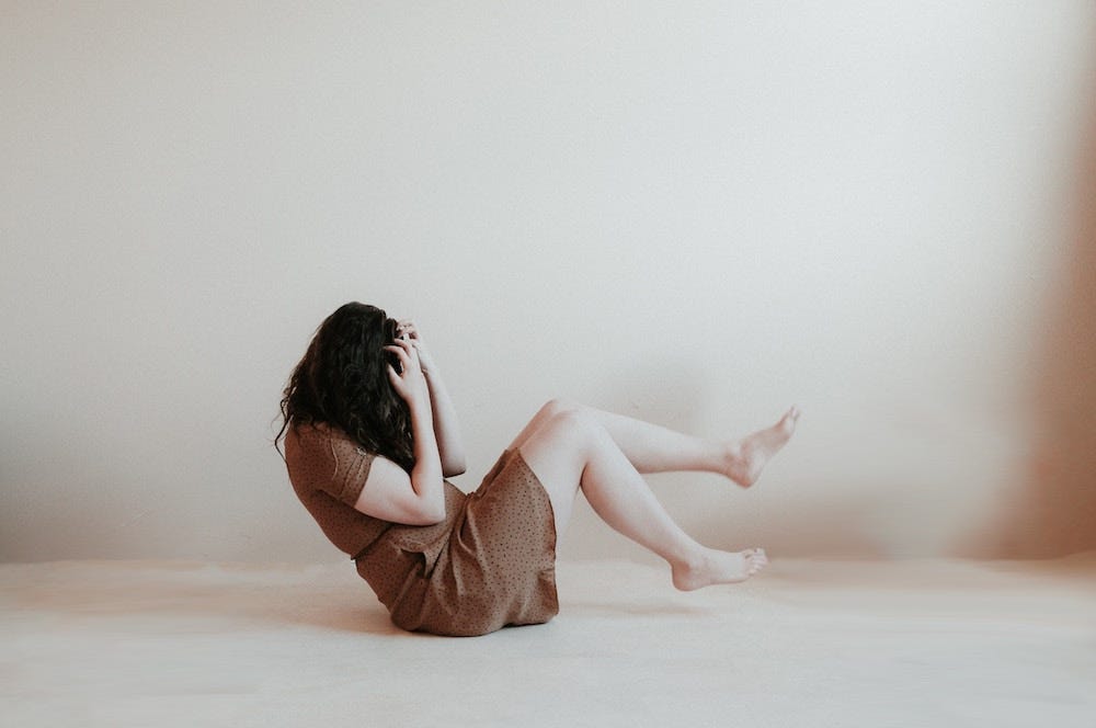 Woman in brown dress with bare feet sitting on the floor covering her face with her hands.
