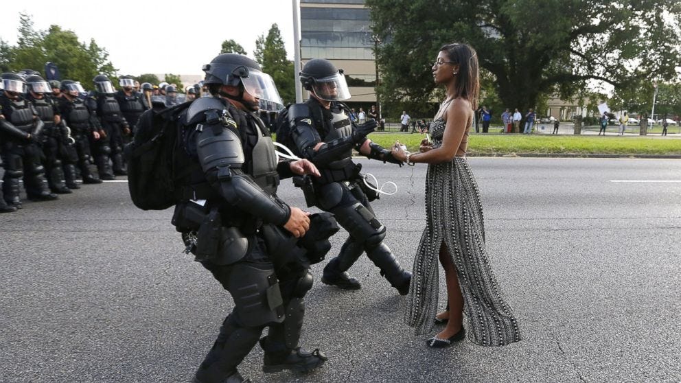 African american lady with black hair and black dress standing still in the middle of the streets while officers in black armors stand against her and rush towards her