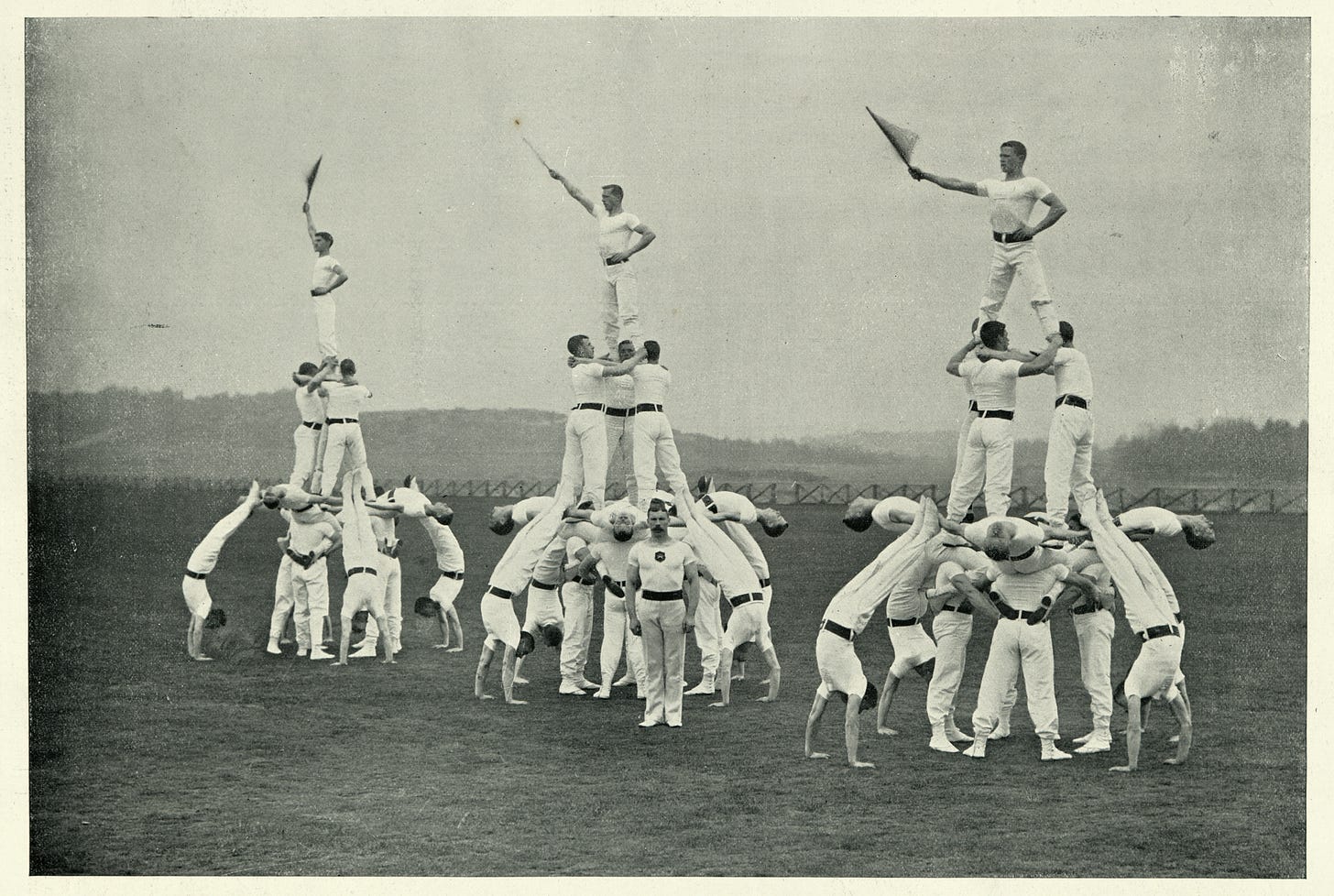 Three groups of about ten men apiece, all dressed in white trousers and T-shirts, form standing human pyramids. The man at the top of each pyramid holds a flag in the air with his right hand, with his left hand placed on his hip. Three groups of about ten men apiece, all dressed in white trousers and T-shirts, form standing human pyramids. The man at the top of each pyramid holds a flag in the air with his right hand, with his left hand placed on his hip.