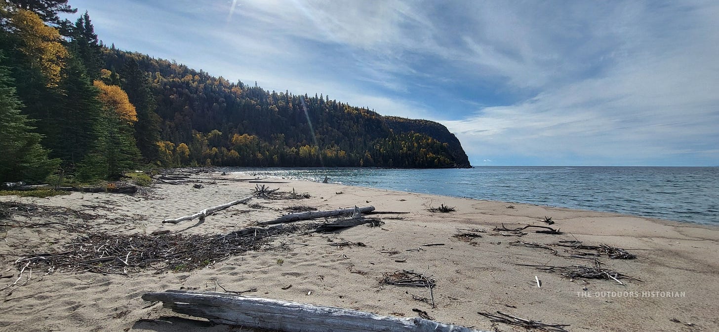 The sheer granite cliffs of Old Woman Bay seen from a sandy beach under a shifting, sunlit grey sky.
