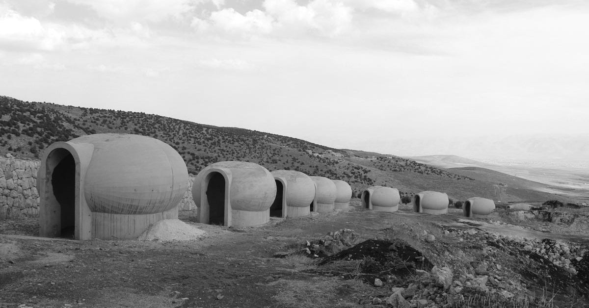 A line of dome-shaped concrete prayer cells stretches across a barren Lebanese hillside under an open sky—each identical, austere, and open to the desert wind. The landscape is dry and silent, evoking monastic discipline and isolation. These structures form the world’s largest rosary installation in Deir El Ahmar, Lebanon—a brutalist litany cast in stone