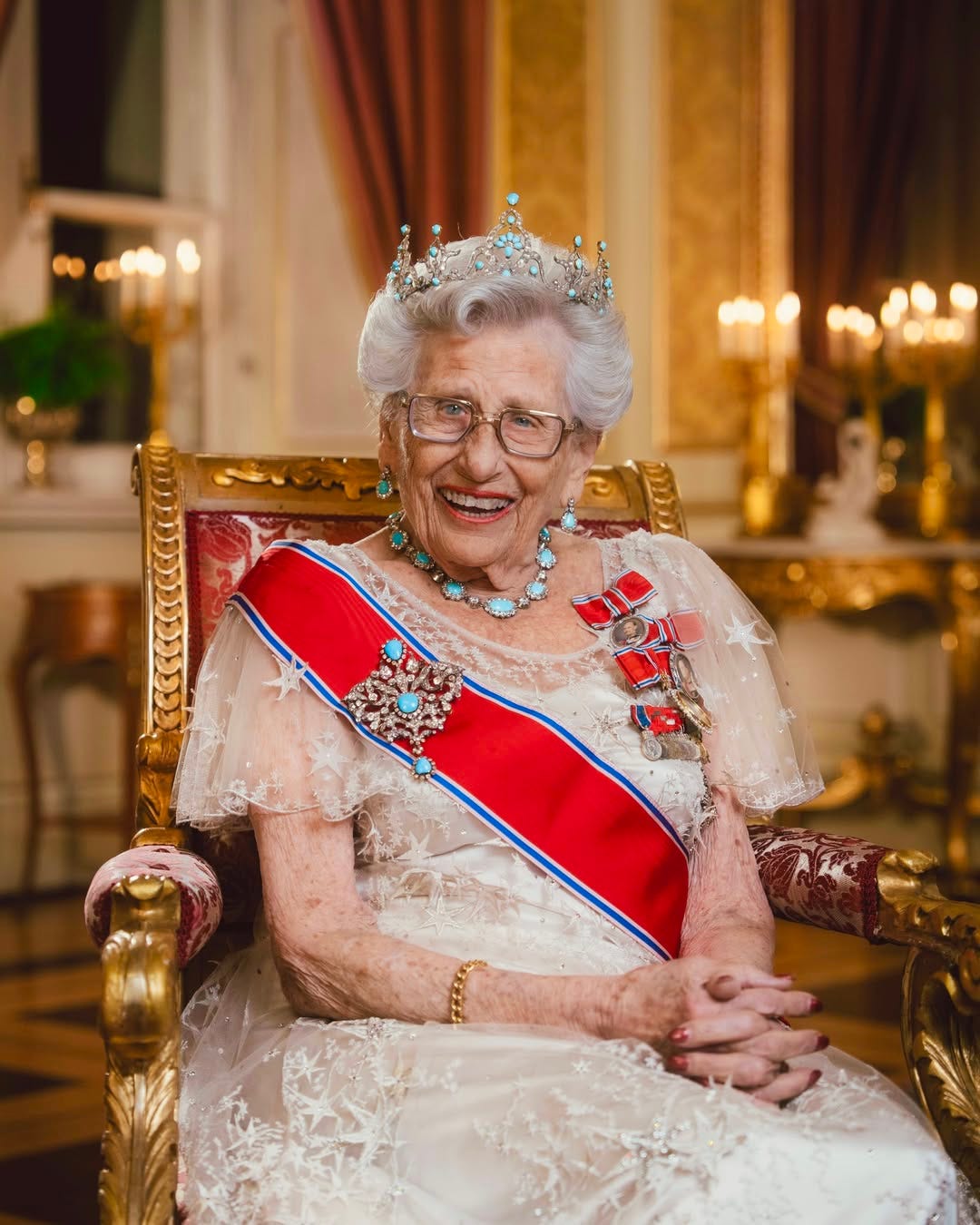 Princess Astrid sat down smiling a white lace dress, red sash and crown