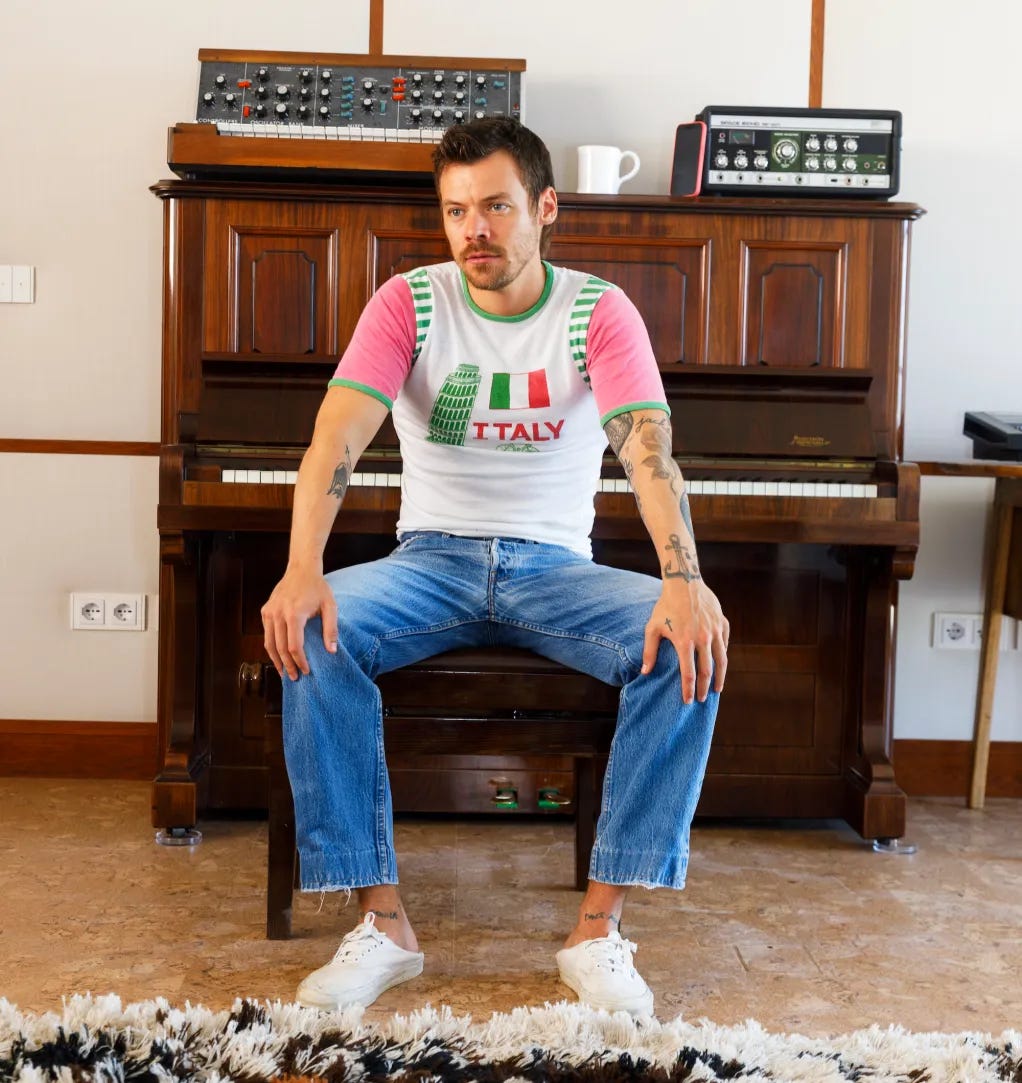 Harry Styles sitting on a piano bench wearing a white t-shirt with an Italian flag and "ITALY" printed on it. Harry Styles sitting on a piano bench wearing a white t-shirt with an Italian flag and "ITALY" printed on it.