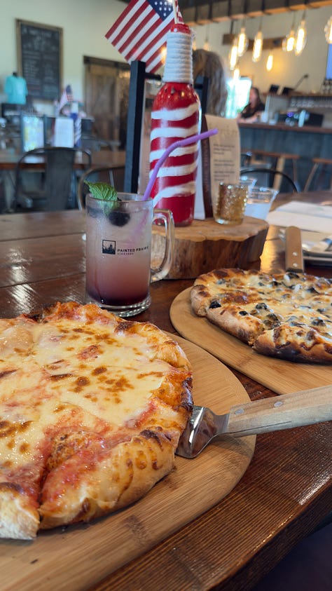 Two very cheesy pizzas on wooden planks and a fizzy drink topped with blackberries in a mug. | A back patio with fire pit, plenty of seating, and a vineyard. | A person holding a bright orange drink in a mug reading “Painted Prairie”