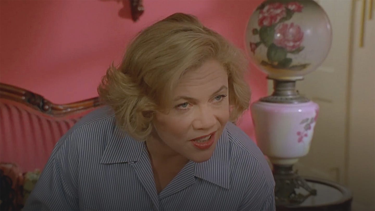 Film still from Serial Mom (1994), showing Kathleen Turner mid-sentence, leaning forward in a blue pinstripe blouse against a hot pink wall and antique furniture. Her expression is mischievous and knowing, as she needle-drops "pussy willows" to her neighbor, Dottie Hinkle. Film still from Serial Mom (1994), showing Kathleen Turner mid-sentence, leaning forward in a blue pinstripe blouse against a hot pink wall and antique furniture. Her expression is mischievous and knowing, as she needle-drops "pussy willows" to her neighbor, Dottie Hinkle.