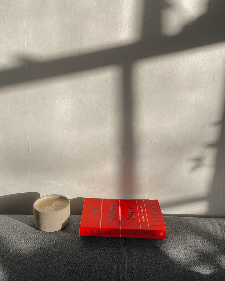 Image 1: The book All About Love and a coffee mug sit on the sofa armrest, with a glimpse of cosy decor in the background. / Image 2: A close-up of the book’s spine and coffee, with soft light and shadows playing on the wall.
