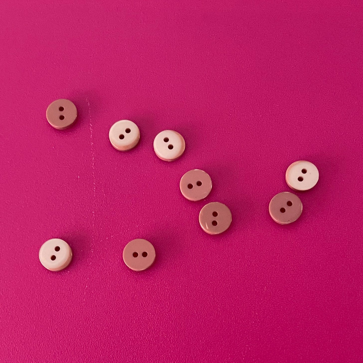 Nine small buttons from a 1980s shirt, scattered across a bright pink surface. The buttons are round, plastic, and two-holed, in shades of beige and muted brown.