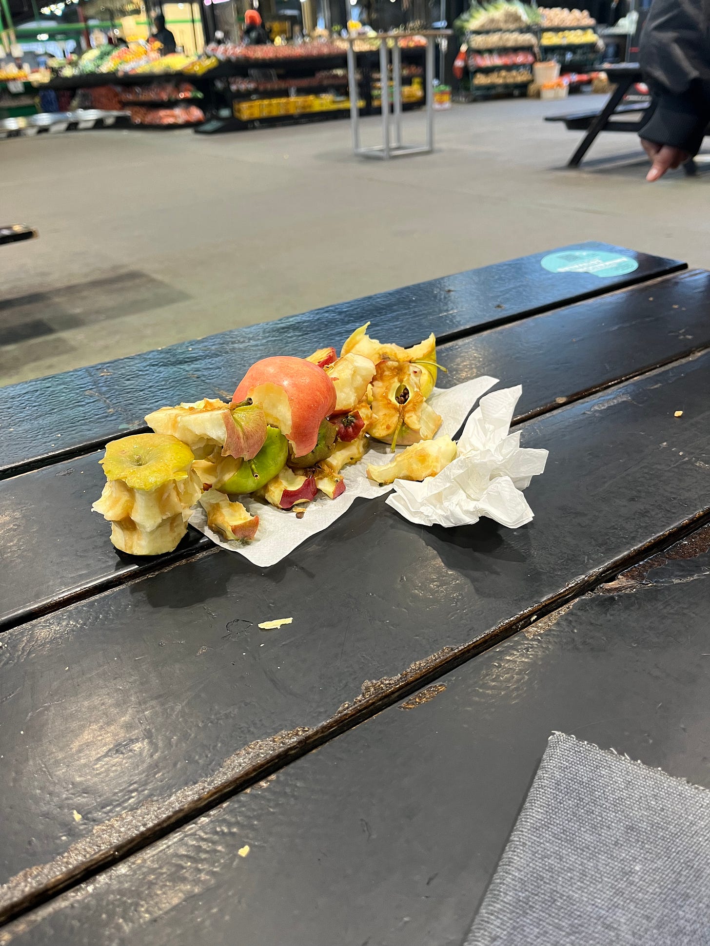 A pile of apple cores sits on a napkin on a black picnic table. Behind the table, blurry fruit stands are visible in the background.