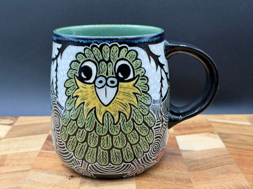 A simply spectacular sgraffito ceramic mug with a bold, charismatic Kākāpō parrot taking up most of the visible space. It has a yellow beard and green feathers.