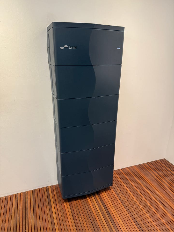 A photo of a vertical, black home battery, and a wide image showing a showroom kitchen with a large screen displaying energy usage statistics.