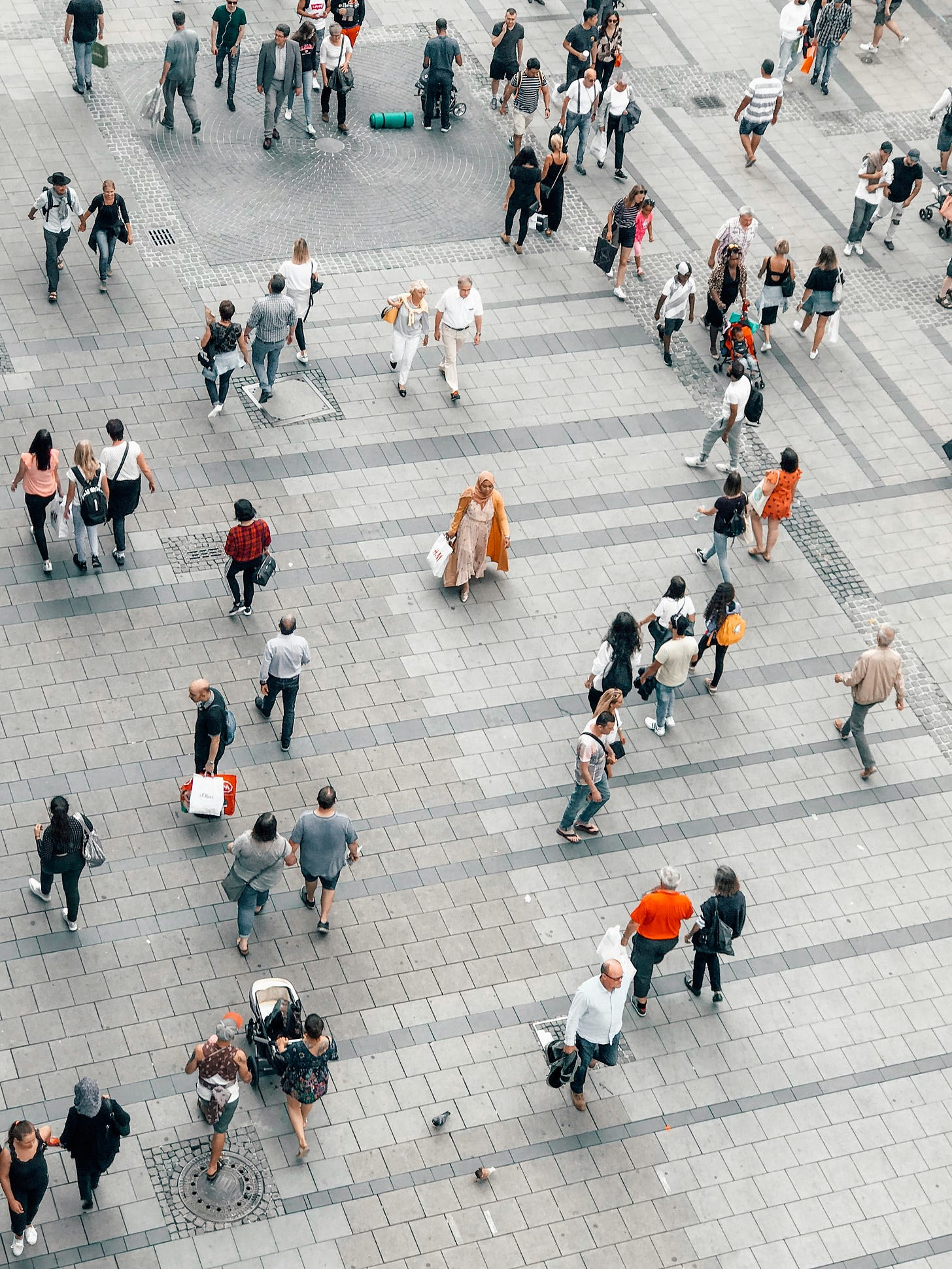 An aerial photo of a grey-paved urban plaza filled with people walking in different directions: shoppers, commuters, tourists, couples, families, friends. An aerial photo of a grey-paved urban plaza filled with people walking in different directions: shoppers, commuters, tourists, couples, families, friends.
