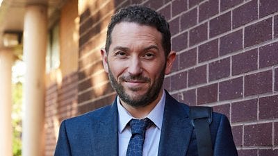 A man in a blue suit and shirt smiles to camera with a bag slung over one shoulder