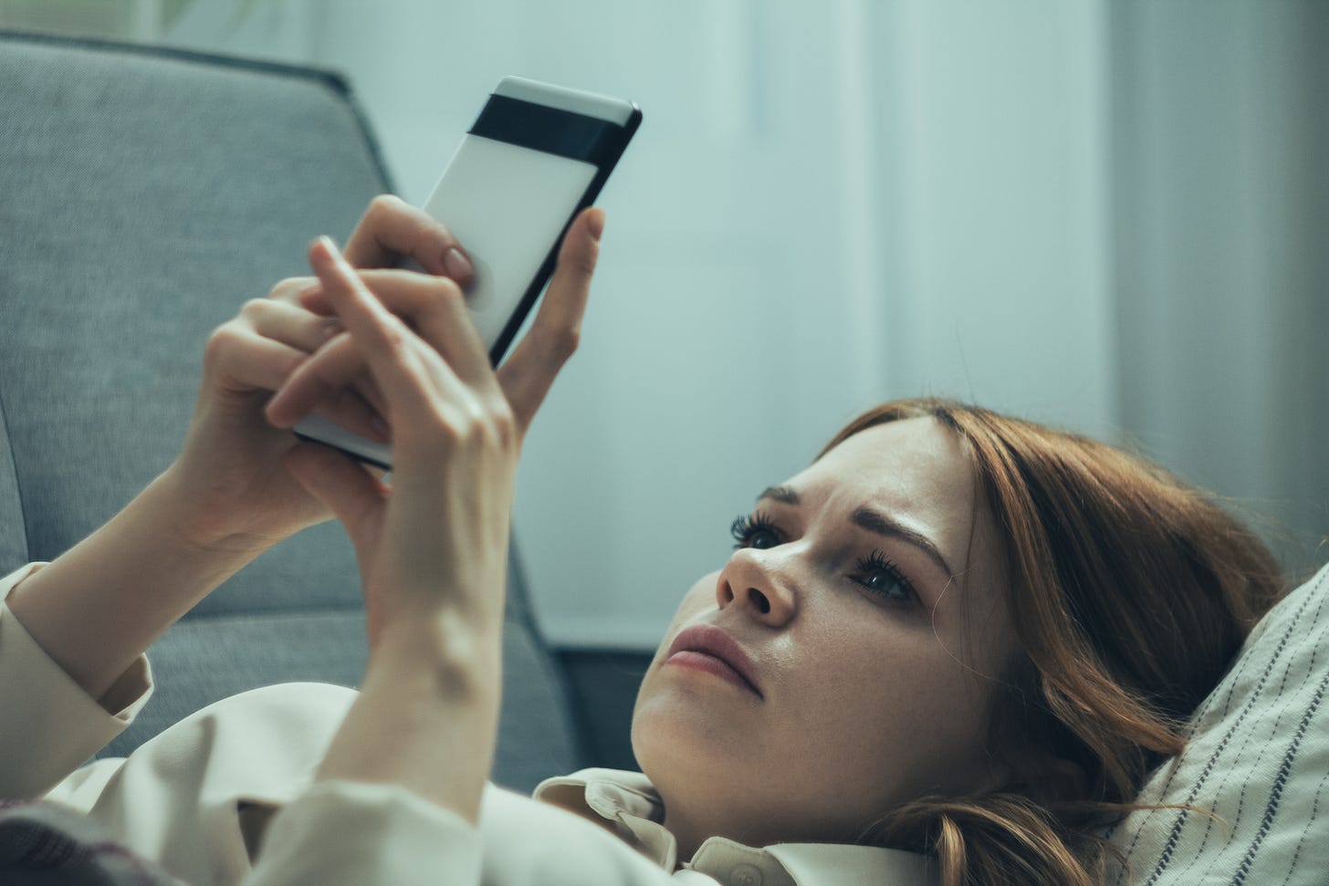 A woman lying on a sofa staring into her phone with a look of fear and panic on her face.