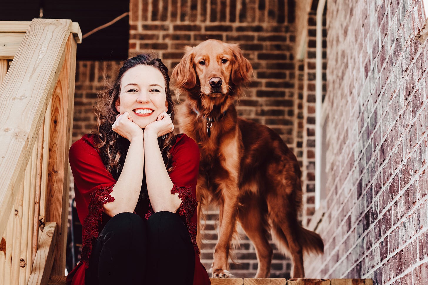 A woman sits on steps smiling at the camera with a golden retriever behind her