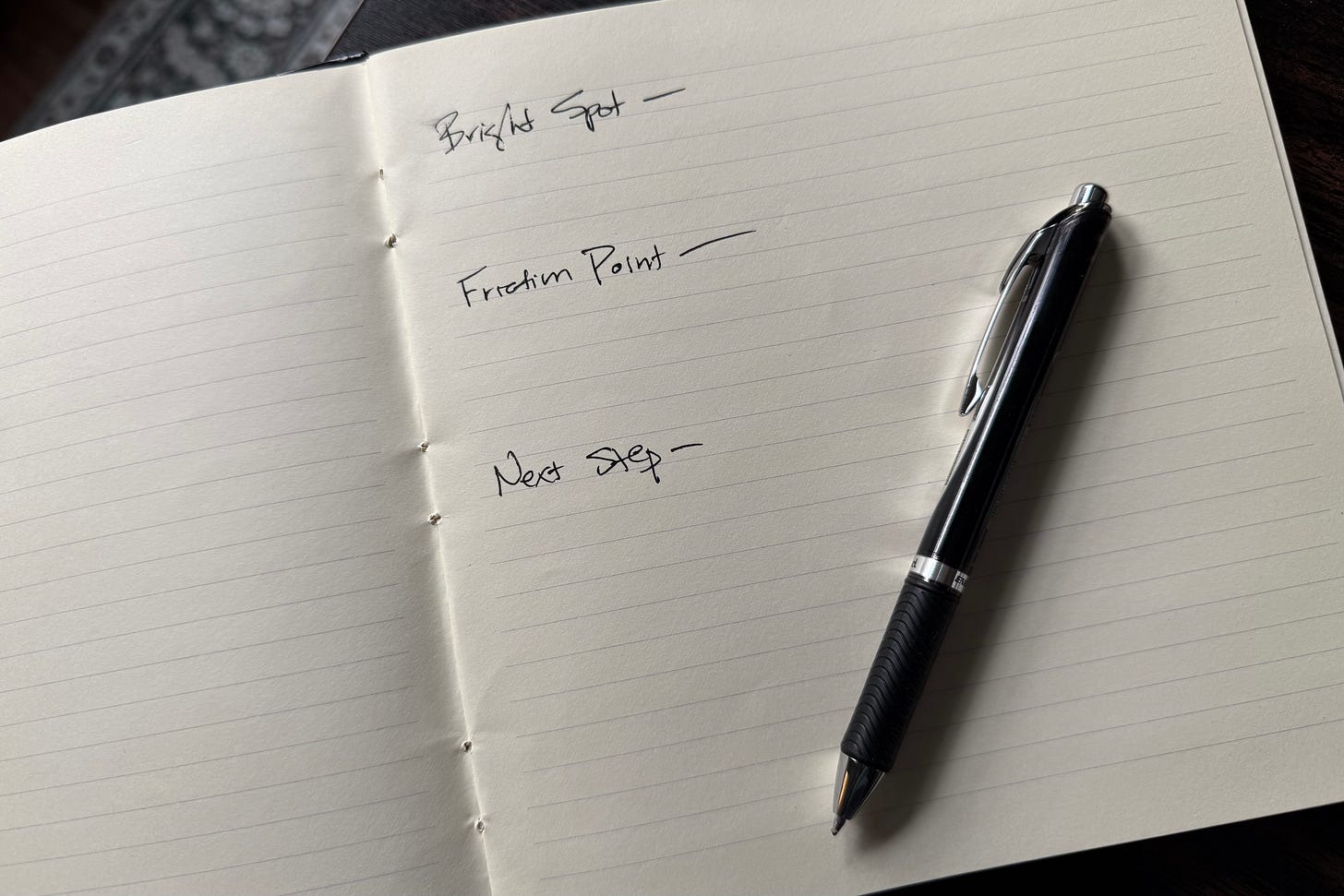 Open notebook with pen; three handwritten prompts—Bright spot, Friction point, Next step.” Open notebook with pen; three handwritten prompts—Bright spot, Friction point, Next step.”