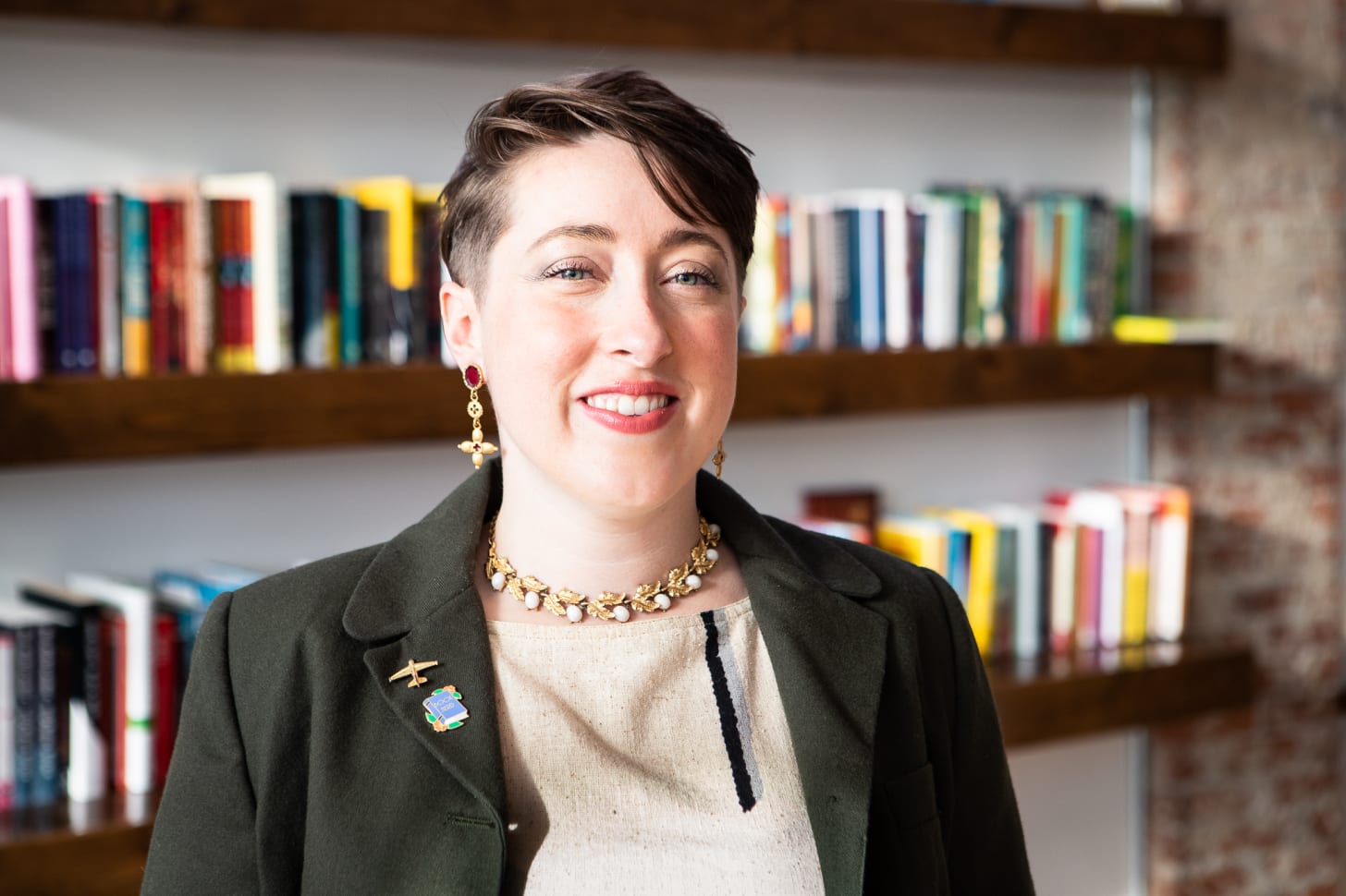 A headshot of Mandy Shunnarah, a pale human with short brown hair and green eyes smiling at the camera while wearing a beige linen shirt with vertical stripes and a green blazer. They are standing in front of a bookshelf full of colorful books A headshot of Mandy Shunnarah, a pale human with short brown hair and green eyes smiling at the camera while wearing a beige linen shirt with vertical stripes and a green blazer. They are standing in front of a bookshelf full of colorful books