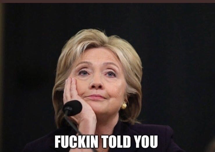 Hillary Clinton with blonde hair faces forward against a dark background, her right hand resting on her cheek in a facepalm gesture, wearing a purple suit jacket and gold earrings, with white text overlay reading FUCKIN TOLD YOU.