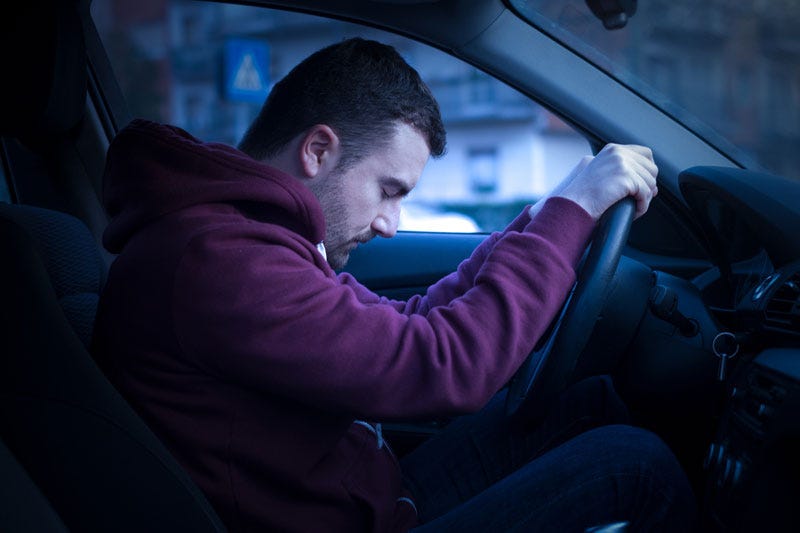 Tired man in maroon hoodie slumped over the steering wheel of a car at night, eyes closed. Tired man in maroon hoodie slumped over the steering wheel of a car at night, eyes closed.