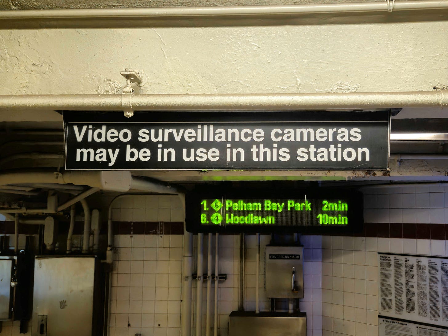 A photograph of an MTA sign in a subway station that says "Video surveillance cameras may be in use in this station." A photograph of an MTA sign in a subway station that says "Video surveillance cameras may be in use in this station."