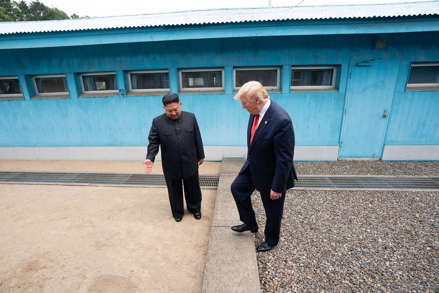 Kim Jong Un gestures while Donald Trump steps over a concrete border marking the separation between two areas outside a blue building.