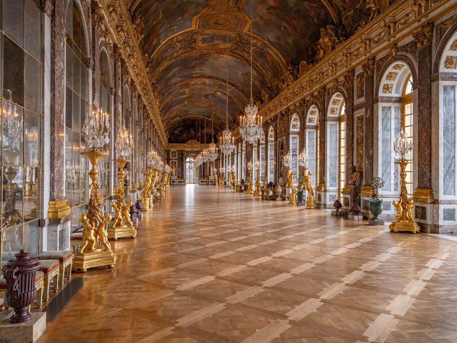 Wide, empty view down the length of the Hall of Mirrors at the Palace of Versailles. The polished marble floor, repeating chandeliers, arched mirrors, and gilded ornament create an endless corridor of reflections. The symmetry and multiplied images evoke themes of power, distortion, and the self-replicating architecture of monarchy. Wide, empty view down the length of the Hall of Mirrors at the Palace of Versailles. The polished marble floor, repeating chandeliers, arched mirrors, and gilded ornament create an endless corridor of reflections. The symmetry and multiplied images evoke themes of power, distortion, and the self-replicating architecture of monarchy.