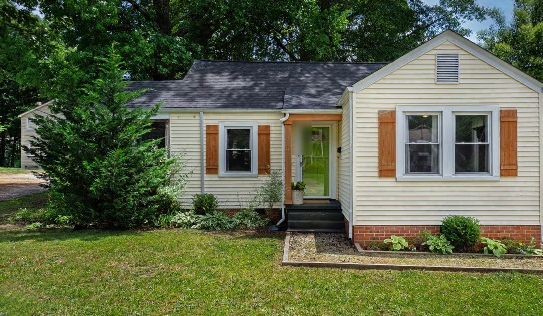 A single-story cottage with pale yellow siding, wooden shutters, a green front door and a brick foundation, behind a lawn with a young evergreen and hostas, under tall shade trees