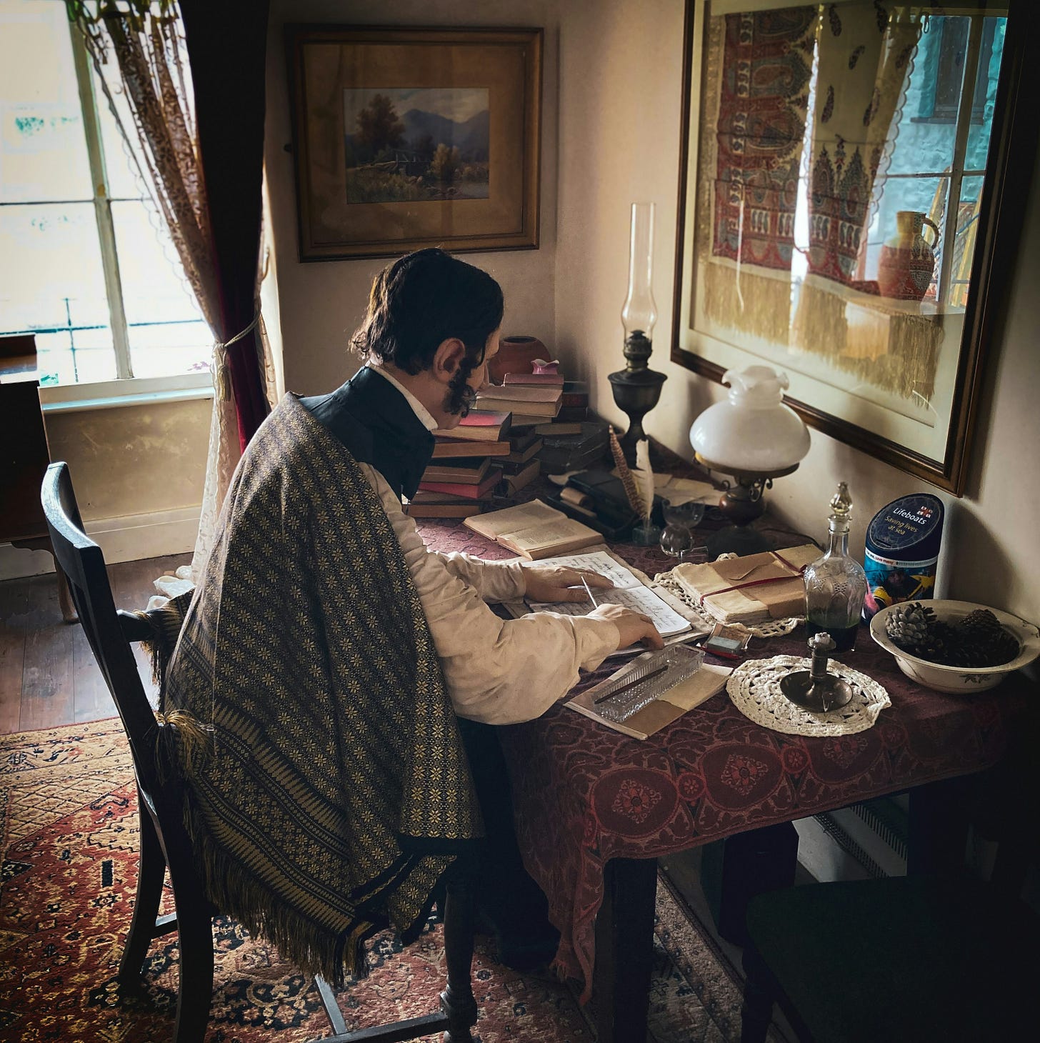 Victorian writer working at desk piled with books and with pictures hanging nearby