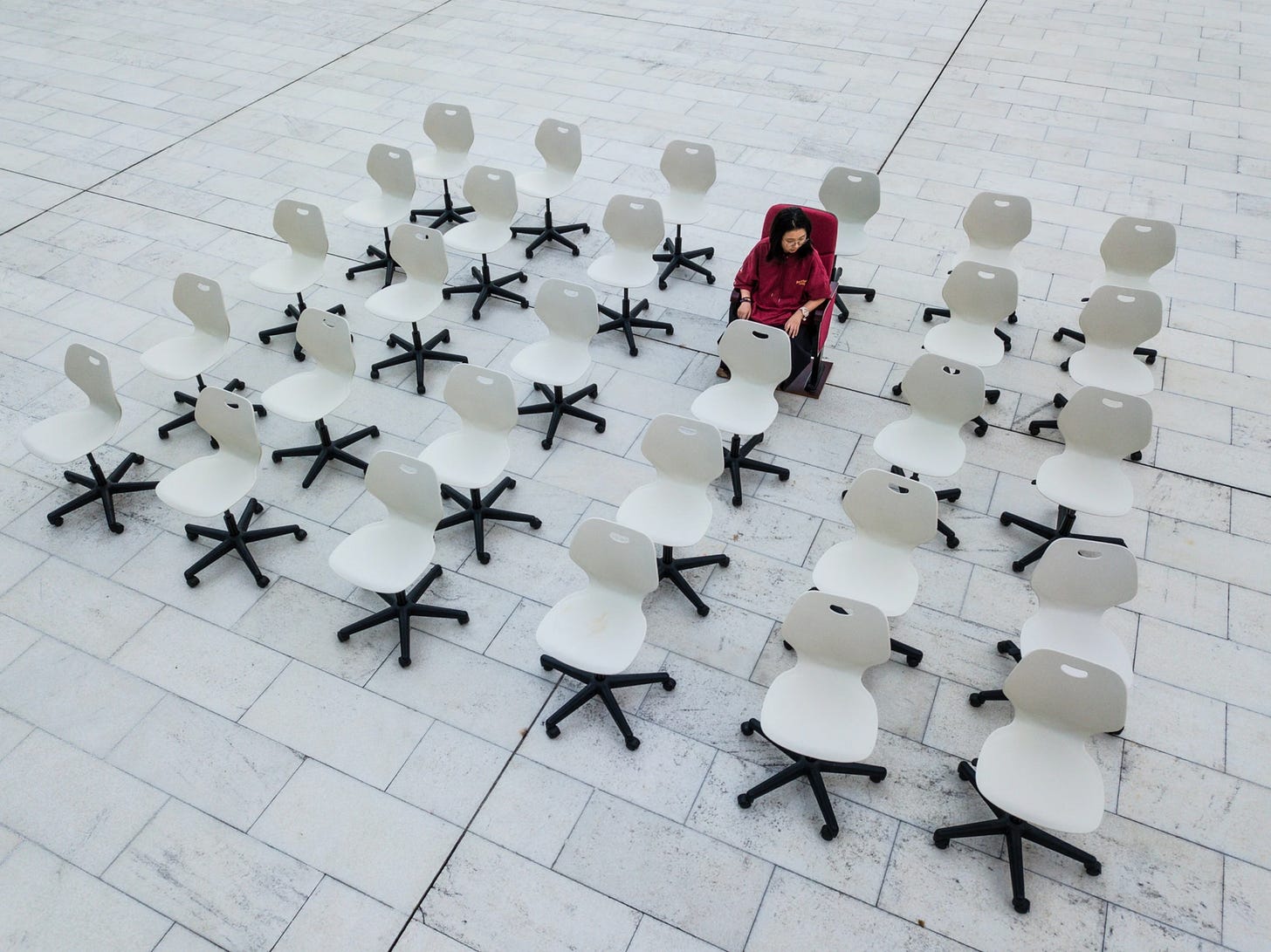 Installation view: An aerial view of a person in a red chair, surrounded by a grid of empty white swivel chairs on a large tiled surface. The person, wearing a red outfit, sits slightly apart from the uniform arrangement, creating a striking visual contrast.