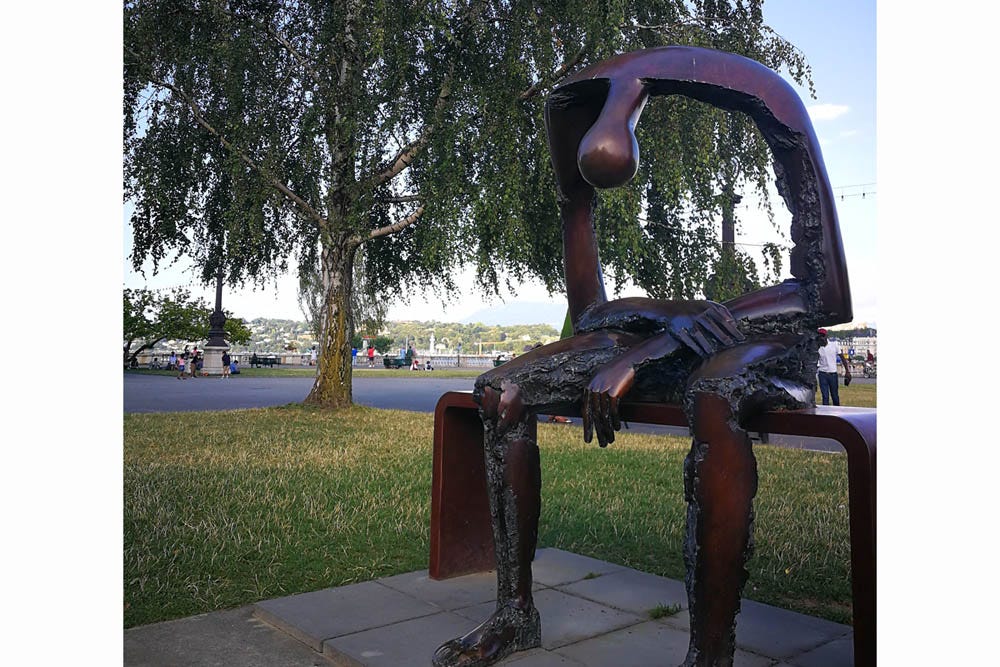 Photo of a copper statue depicting an abstract human figure seated on a bench, its featureless head hanging down from its hunched shoulders, its lower arms loosely crossed in its lap. The upper arms alone support the shoulders, neck, and head, leaving the figure with no torso between the waist and shoulders, only negative space suggesting the emptiness of loss and grieving. Portions of the arms and legs are in rough, unpolished metal, suggesting that the figure is decaying as it sits. Photo of a copper statue depicting an abstract human figure seated on a bench, its featureless head hanging down from its hunched shoulders, its lower arms loosely crossed in its lap. The upper arms alone support the shoulders, neck, and head, leaving the figure with no torso between the waist and shoulders, only negative space suggesting the emptiness of loss and grieving. Portions of the arms and legs are in rough, unpolished metal, suggesting that the figure is decaying as it sits.