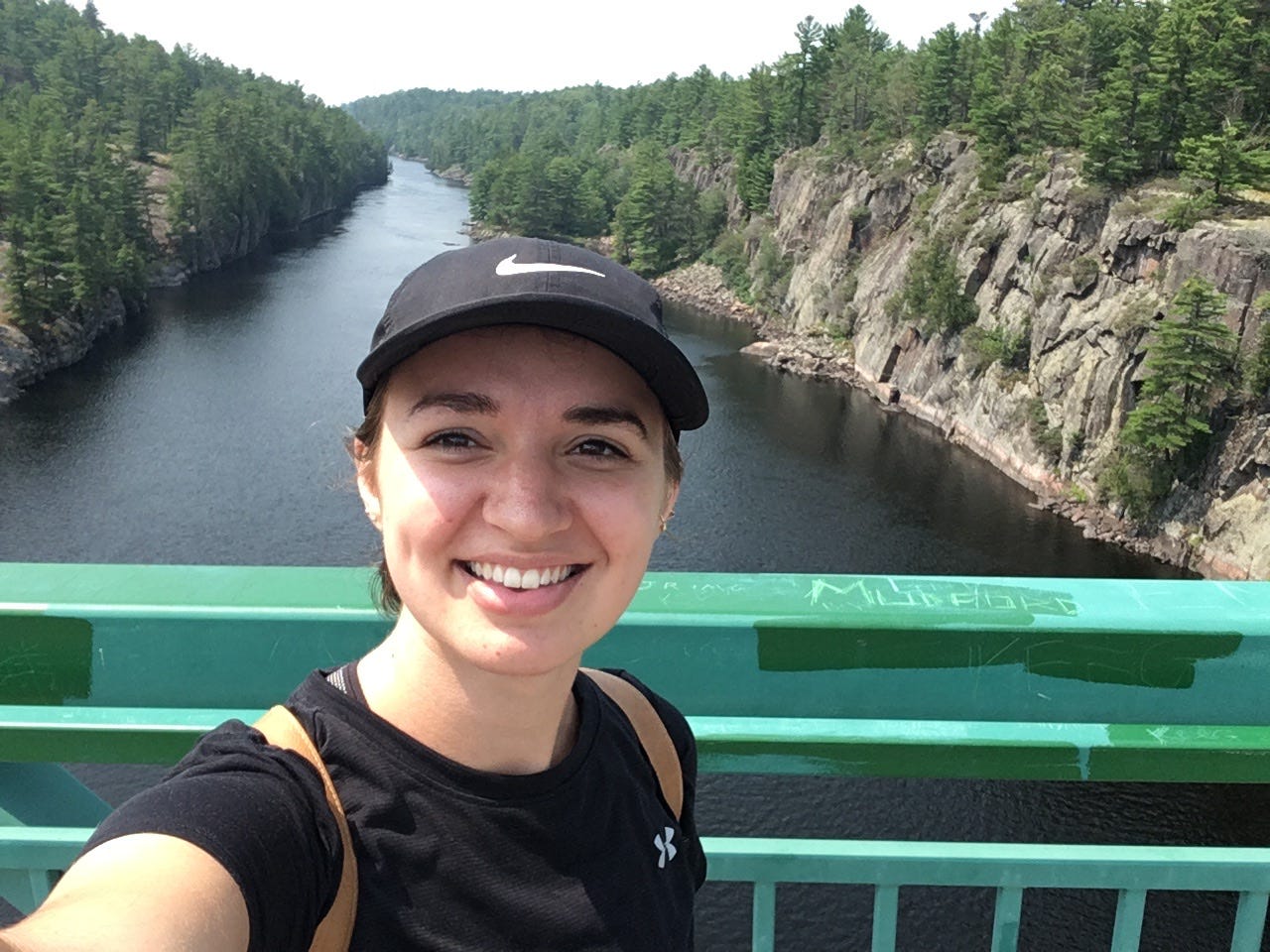 Close-up selfie of Sadie smiling on a green bridge with a river and forest in the background. Close-up selfie of Sadie smiling on a green bridge with a river and forest in the background.